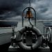 A grayscale shot of a lifesaver ring and a golden bell on a boat sailing the ocean during a mischievous stormy weather